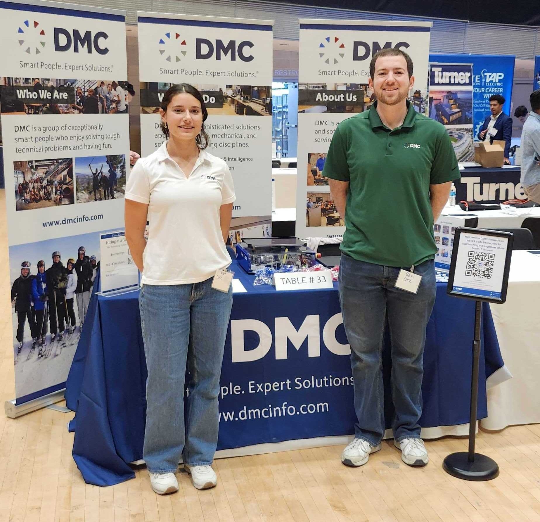 Two DMC team members standing in front of DMC's booth at a career fair.
